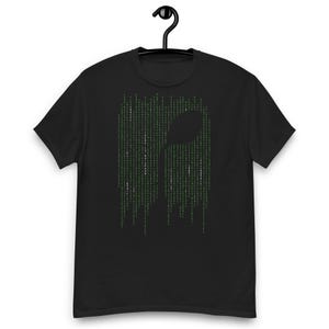 Puede incluir: Una camiseta negra de manga corta con un gráfico central de la silueta de una cuchara formada por columnas verticales de código digital verde. El texto "THERE IS NO SPOON" está integrado en blanco en el patrón de código, creando un diseño.