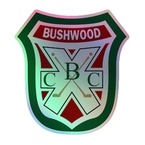 May include: A shield-shaped sticker with a holographic finish featuring a dark green border and a red background. The text BUSHWOOD is printed at the top. A white cross design contains the letters C, B, and C with two crossed golf clubs.