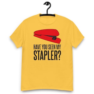 May include: Mustard yellow t-shirt featuring a red stapler graphic and the text "HAVE YOU SEEN MY STAPLER?" in black. The shirt is suspended from a black hanger.
