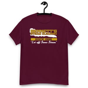 Pode incluir: Uma camiseta bordô pendurada em um cabide preto. A camiseta apresenta o texto "BENNETT'S STEAM CLEANING SINCE 1985" em dourado e branco, com a frase "Let off Some Steam" abaixo.