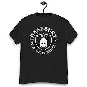 Può includere: T-shirt nero con grafica bianca. Il design include il testo "DANEBURY METAL DETECTING CLUB" attorno a uno scudo con "D.M.D.C" e un elmo. La maglietta presenta anche "Estd. 1978".
