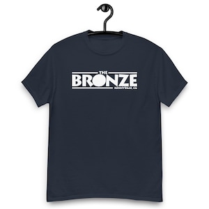 Könnte beinhalten: Marineblaues T-Shirt mit dem weißen Text "THE BRONZE" in einem rechteckigen Design. Darunter steht "SUNNYVALE, CA". Das T-Shirt hängt an einem schwarzen Kleiderbügel.
