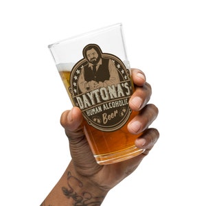 Könnte beinhalten: Ein klares Glasbierkrug mit einem braunen und weißen Aufkleber, der einen Cartoon-Mann mit Bart und den Text "Daytona's Human Alcoholic Beer" zeigt.