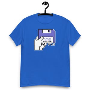 以下が含まれることがあります： 明るいブルーの半袖Tシャツで、手でライトパープルのフロッピーディスクを持っているグラフィックが描かれています。ディスクのラベルには黒い文字で「AMIGA Work bench V 1.3」と書かれています。グラフィックはハンガーに掛けられたTシャツの胸の中央に配置されています。