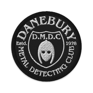 May include: A black and white embroidered patch with the text "DANEBURY METAL DETECTING CLUB" encircling a shield design. The shield features a helmet graphic and the text "D.M.D.C." and "1978".