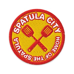 May include: Embroidered patch with a yellow center and a red border. The patch features the text "SPATULA CITY HOME OF THE SPATULA" in white, surrounding two crossed red spatulas.