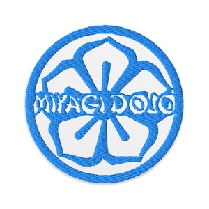 Peut inclure: Écusson brodé circulaire avec un motif floral bleu et le texte blanc "MIYAGI DOJO". L'écusson présente un aspect détaillé et cousu, adapté à l'application sur des vêtements ou des accessoires. Le design est sur fond blanc.
