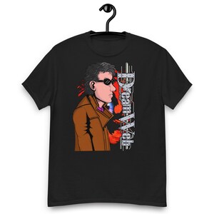 Może przedstawiać: Czarny t-shirt z krótkim rękawem z nadrukiem graficznym przedstawiającym osobę w brązowym płaszczu i ciemnych okularach przeciwsłonecznych. Obok postaci widnieje pionowy napis "Dream Web" wykonany metaliczną czcionką na czerwonym tle.