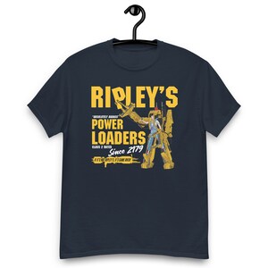 May include: A navy blue t-shirt with a yellow graphic of a cartoon character operating a construction vehicle. The graphic includes the text "RIPLEY'S", "ABSOLUTELY BADASS", "POWER LOADERS", "CLASS 2 RATED", "Since 2179", and "IF IT'S NOT RIPLEY'S IT'S GAME OVER".