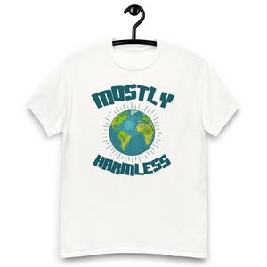 Könnte beinhalten: Weißes T-Shirt mit dem Text "MOSTLY HARMLESS" in türkisfarbenen Buchstaben. Eine Weltkugel-Grafik in Türkis und Gelb befindet sich zwischen den Wörtern. Das T-Shirt hängt an einem schwarzen Kleiderbügel.