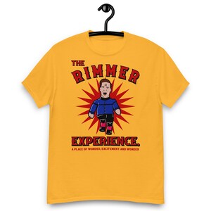 May include: Mustard yellow t-shirt featuring a cartoon illustration of a man in a blue jacket and pink boots, set against a red burst design. The text "THE RIMMER EXPERIENCE" is above and below the illustration, with the tagline "A PLACE OF WONDER, EXCITEMENT AND WONDER."