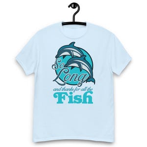 Könnte beinhalten: Hellblaues T-Shirt mit einer Grafik von zwei Delfinen in einem blauen Kreis, mit dem Text "So Long and thanks for all the Fish" in Weiß und Türkis. Das T-Shirt hängt an einem schwarzen Kleiderbügel.