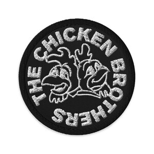 May include: A round, black embroidered patch with the text "THE CHICKEN BROTHERS" in white. The patch features a white outline of two cartoon chickens facing each other. The patch is likely for clothing or accessories.