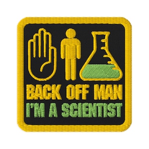 Pode incluir: Patch bordado com fundo preto e borda amarela. O patch apresenta desenhos de linha amarelos de uma mão, uma figura de palito e um balão com líquido verde. O texto diz "BACK OFF MAN I'M A SCIENTIST" em amarelo e verde.