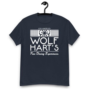 May include: A navy blue short-sleeve t-shirt featuring a white graphic print. The design includes the text "LOS ANGELES", "WOLF HART'S", and "Fine Dining Experiences" centered above a stylized floral icon enclosed within a rectangular border.