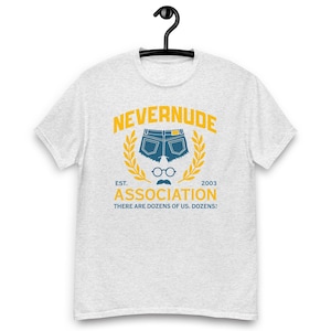May include: A light gray t-shirt with a graphic design featuring the words "NEVERNUDE ASSOCIATION" in yellow, with a pair of blue jean shorts, glasses, and a mustache. The text "THERE ARE DOZENS OF US. DOZENS!" is below.