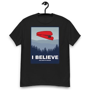 May include: Black t-shirt featuring a red stapler graphic above a mountain landscape. The text "I BELIEVE" and "YOU HAVE MY STAPLER" are printed below. The shirt is suspended from a black hanger.