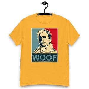 May include: Mustard yellow t-shirt featuring a stylized portrait with the word "WOOF" below. The portrait has a blue and red background. The design is printed on a short-sleeved, crew neck t-shirt.