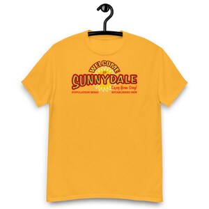 May include: Mustard yellow t-shirt featuring the text "Welcome to Sunnydale" in red and white. The shirt also includes the text "Enjoy Your Stay!" and "Population 38500 Established 1909". The t-shirt is hanging on a black hanger.