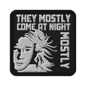 Peut inclure: Écusson brodé noir avec le texte blanc « THEY MOSTLY COME AT NIGHT » et « MOSTLY ». Un visage blanc avec des cheveux flottants est sur le côté gauche de l'écusson.