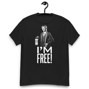 May include: Black t-shirt featuring a black and white image of a man holding a cage, with the text "I'M FREE!" in white block letters. The man is wearing a suit and suspenders. The shirt is hanging on a black hanger.