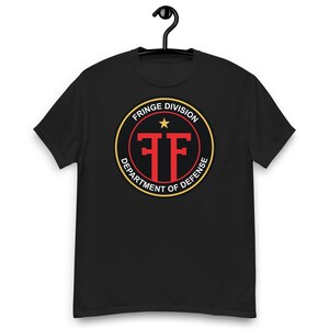 May include: Black t-shirt with a circular graphic featuring the text "FRINGE DIVISION DEPARTMENT OF DEFENSE". The graphic is red, yellow, and white. The shirt is hanging from a black hanger.