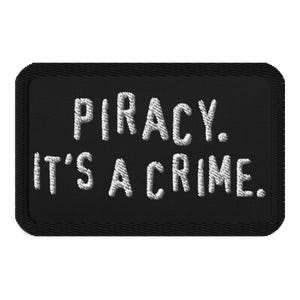 May include: A black rectangular patch with white embroidered text that reads "PIRACY. IT'S A CRIME." The patch has a stitched border.