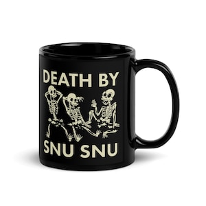 Op de afbeelding: Een zwarte keramische mok met een afbeelding van drie gebroken witte skeletten in verschillende houdingen. De tekst "DEATH BY SNU SNU" is in gebroken witte hoofdletters boven en onder de skeletillustratie op de voorzijde van de mok gedrukt.