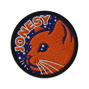 Puede incluir: Parche bordado con el perfil de un gato naranja y bigotes blancos, sobre un fondo azul oscuro con puntos blancos. La palabra "JONESY" está arqueada sobre el gato en letras blancas. El parche tiene un borde negro.