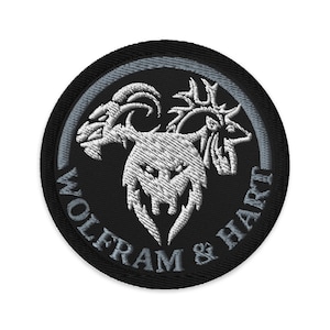 Op de afbeelding: Geborduurde patch met een zwarte achtergrond en een wit en grijs ontwerp. Het ontwerp toont een driekoppig wezen met een ram, hert en wolf. De woorden "WOLFRAM & HART" zijn in het wit rond de rand geborduurd.