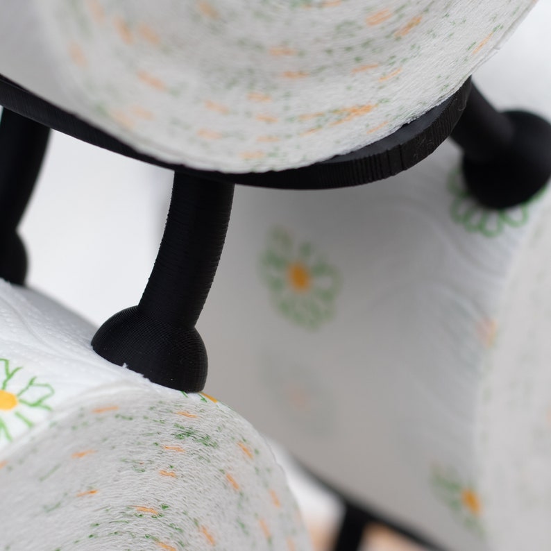 Op de afbeelding: Close-up van een zwarte metalen toiletpapierhouder met twee rollen toiletpapier. Het toiletpapier heeft een witte achtergrond met een groen en geel madeliefjespatroon.