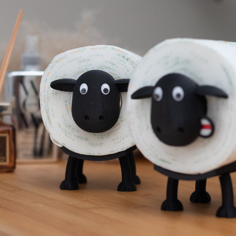 Op de afbeelding: Twee zwart-witte toiletpapierhouders in de vorm van schapen. De schapen hebben zwarte gezichten met witte ogen en een zwarte neus. De schapen staan op een houten oppervlak.