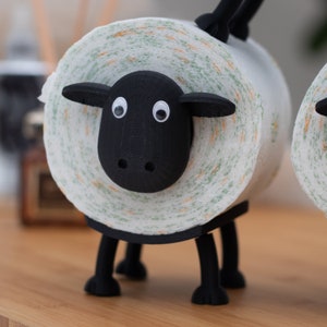 Op de afbeelding: Een zwarte toiletpapierhouder in de vorm van een schaap met een witte en groene gepatroneerde toiletpapierrol. Het schaap heeft zwarte ogen en een zwarte neus.