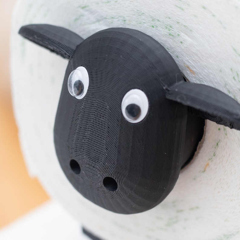 Op de afbeelding: Een zwarte schapenvormige toiletpapierhouder met oogjes. De houder is gemaakt van 3D-geprint plastic en is ontworpen om een standaard rol toiletpapier te houden.