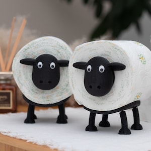 Op de afbeelding: Twee zwart-witte toiletpapierhouders in de vorm van schapen. De schapen zijn gemaakt van wit papier met groene en gele bloemenpatronen. De schapen hebben zwarte gezichten met witte ogen en zwarte neuzen.