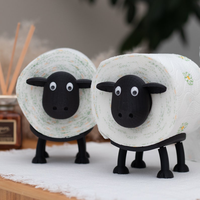Op de afbeelding: Twee zwart-witte toiletpapierhouders in de vorm van schapen. De schapen zijn gemaakt van wit papier met groene en gele bloemenpatronen. De schapen hebben zwarte gezichten met witte ogen en zwarte neuzen.