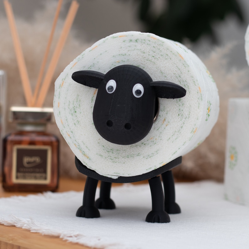 Op de afbeelding: Een zwarte toiletpapierhouder in de vorm van een schaap met een witte rol toiletpapier. Het schaap heeft zwarte ogen en een zwarte neus.
