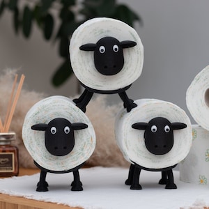 Op de afbeelding: Drie witte toiletrolhouders in de vorm van schapen met zwarte gezichten en poten. De schapen zijn op elkaar gestapeld.
