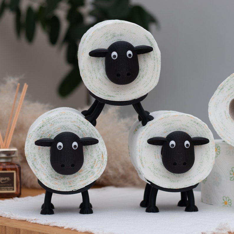 Op de afbeelding: Drie witte toiletrolhouders in de vorm van schapen met zwarte gezichten en poten. De schapen zijn op elkaar gestapeld.