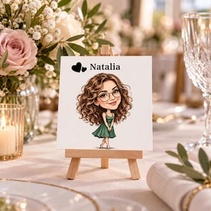 May include: A square white card displayed on a small wooden easel featuring a cartoon illustration of a person with long curly brown hair and glasses wearing a green dress. The text Natalia is printed in black above the illustration with two hearts.