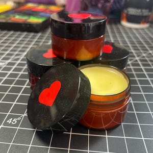 May include: A collection of small, round jars with black lids and red heart accents. The jars are filled with a yellow balm. The jars are stacked on a black grid surface.