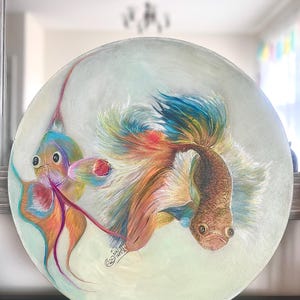 May include: A round, white plate with a painting of two colorful fish. The fish have long, flowing fins and tails. The plate is decorated with a light green background and a signature in the bottom right corner.