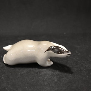 May include: A ceramic badger figurine in a crouching position. The figurine is painted in shades of gray and black, with a white stripe on the face.