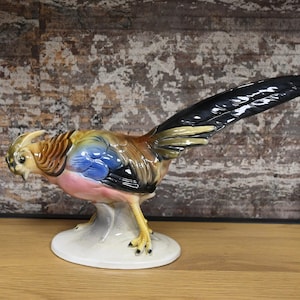 Op de afbeelding: Een keramiek beeldje van een fazant met bruin, goud, blauw en roze verenkleed. De fazant staat op een witte voet.