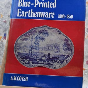 Puede incluir: Un libro azul titulado "Blue-Printed Earthenware 1800-1850" de A.W. Coysh. La portada presenta un plato de cerámica azul y blanco con una escena de paisaje.