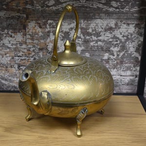 Pode incluir: Uma chaleira de metal dourado com um design ornamentado e uma alça curva. A chaleira tem três pernas e uma tampa com um botão.