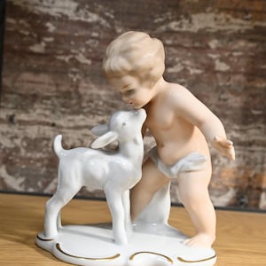 Könnte beinhalten: Eine Porzellanfigur eines Kindes mit blonden Haaren, das ein weißes Rehkitz küsst. Die Figur steht auf einem weißen Sockel mit goldenen Verzierungen.