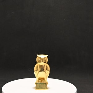 Puede incluir: Una figurita de búho de color dorado con ojos y alas detalladas. El búho está de pie sobre una base circular blanca.