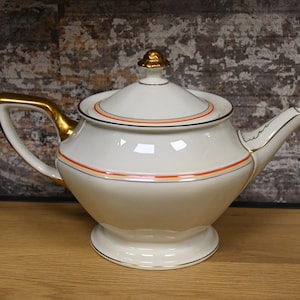Może przedstawiać: Biały czajnik z pozłacanymi zdobieniami i pomarańczowymi i niebieskimi akcentami. Czajnik ma okrągły korpus i pokrywkę z gałką na górze.