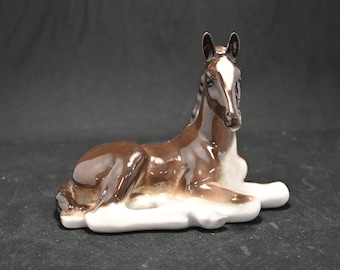 May include: A brown and white porcelain figurine of a lying down foal. The foal has a white stripe down its face and white markings on its legs.
